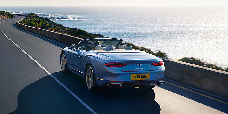 Bentley Marbella New Bentley Continental GTC Azure convertible rear view in Light Windsor blue paint with 22 inch ten spoke wheels driving dynamically on an ocean road in the sunshine