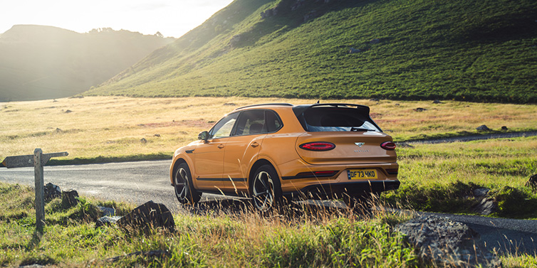 Bentley Marbella Bentley Bentayga S SUV rear three quarter in Sunburst Gold paint on a road surrounded by green hills