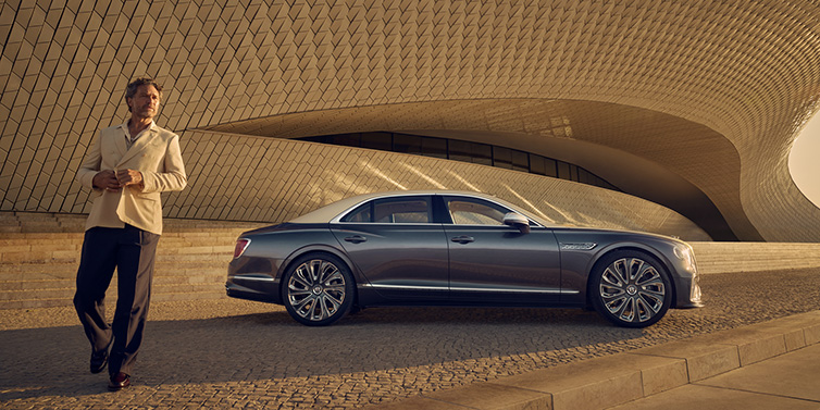 Bentley Marbella Bentley Flying Spur Mulliner sedan in Rose Gold over Magnetic duotone paint parked by a curved wall with person in suit jacket standing on the left